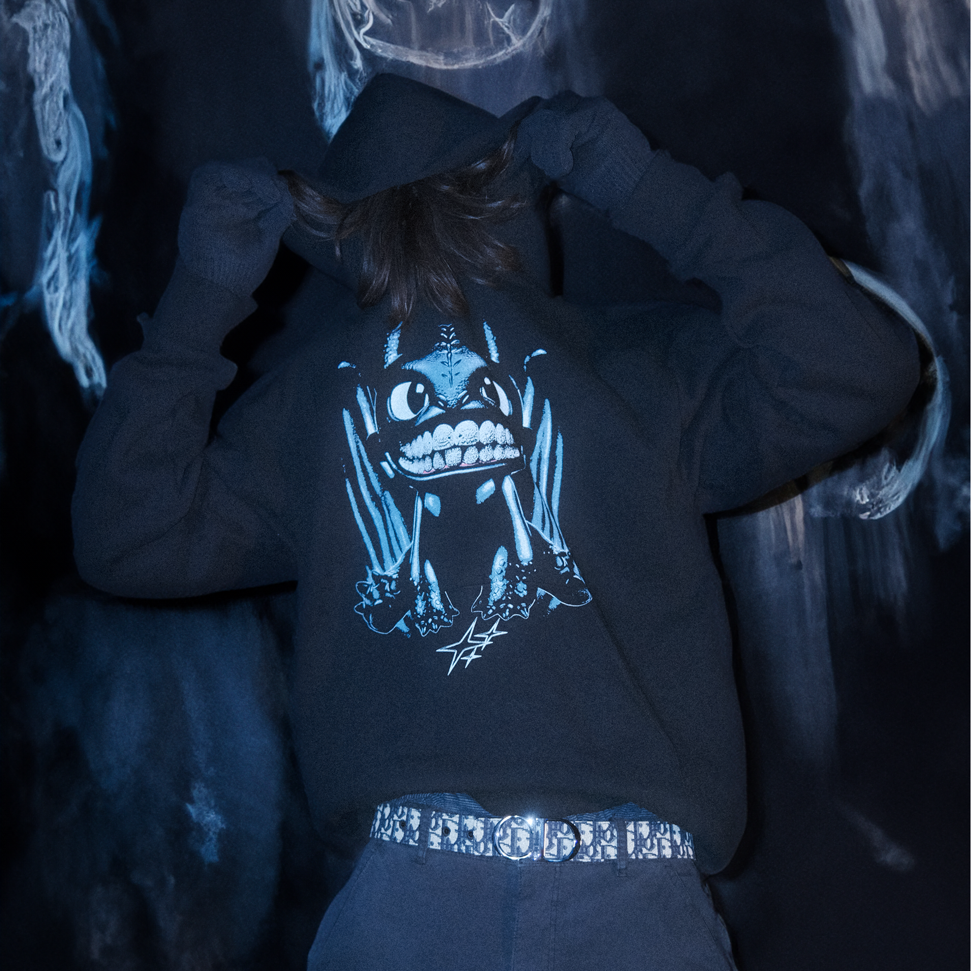 Model wearing a black hoodie with a graphic of a blue dragon character with diamond grillz, sitting over three stars.The model is pulling his hoodie up over his face while standing in a dream like graffitti room.