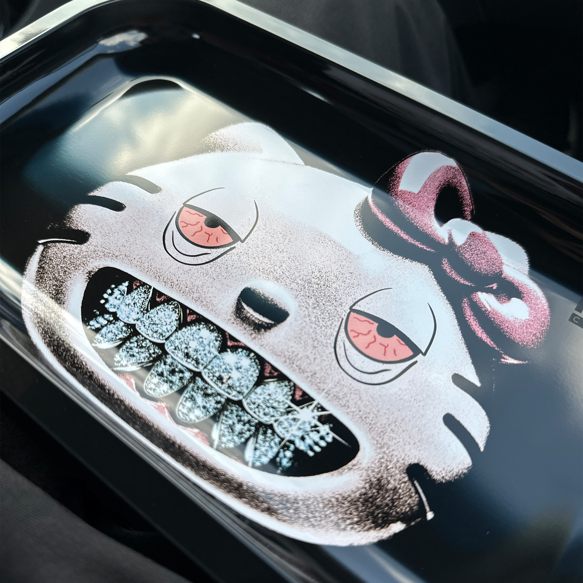 Close up shot of a black rolling tray with a graphic printed showing a light pink cat with a dark pink bow on the left side of its head, with blue diamond grillz and droopy red eyes. There is a logo that shows "noir complete" on the right of the tray in matte black.