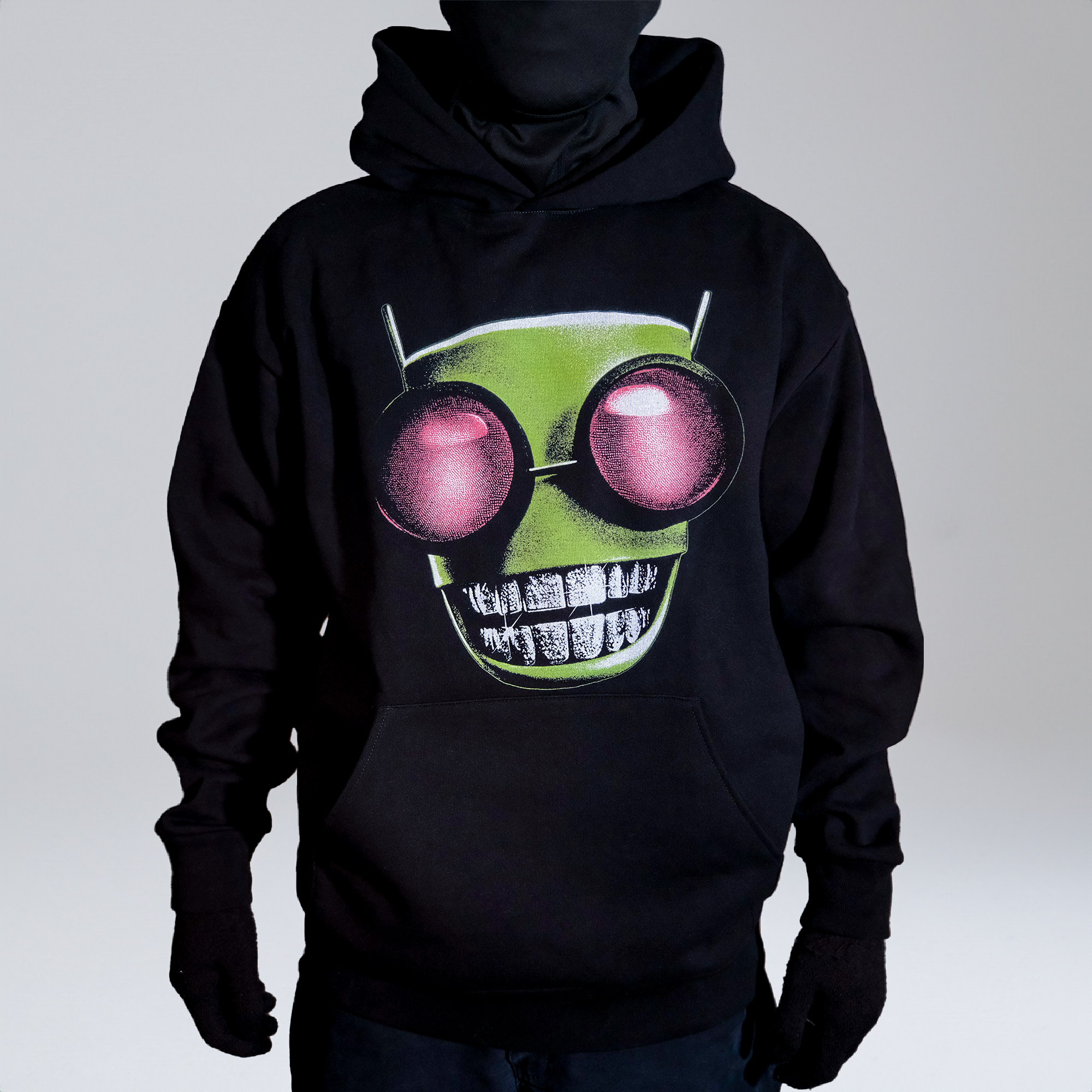 Model wearing a black hoodie with a graphic of a green alien head featuring large red eyes and diamond grillz, standing against a neutral background.