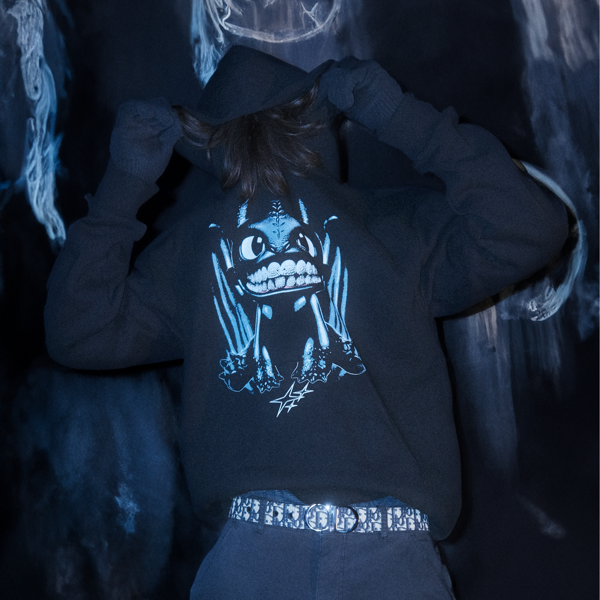 Model wearing a black hoodie with a graphic of a blue dragon character with diamond grillz, sitting over three  stars.The model is pulling his hoodie up over his face while standing in a dream like graffitti room.