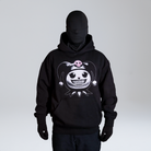 Model wearing a black hoodie featuring a graphic of a stylized character head with pigtails and a pink skull motif, complete with diamond grillz. Standing against a neutral background