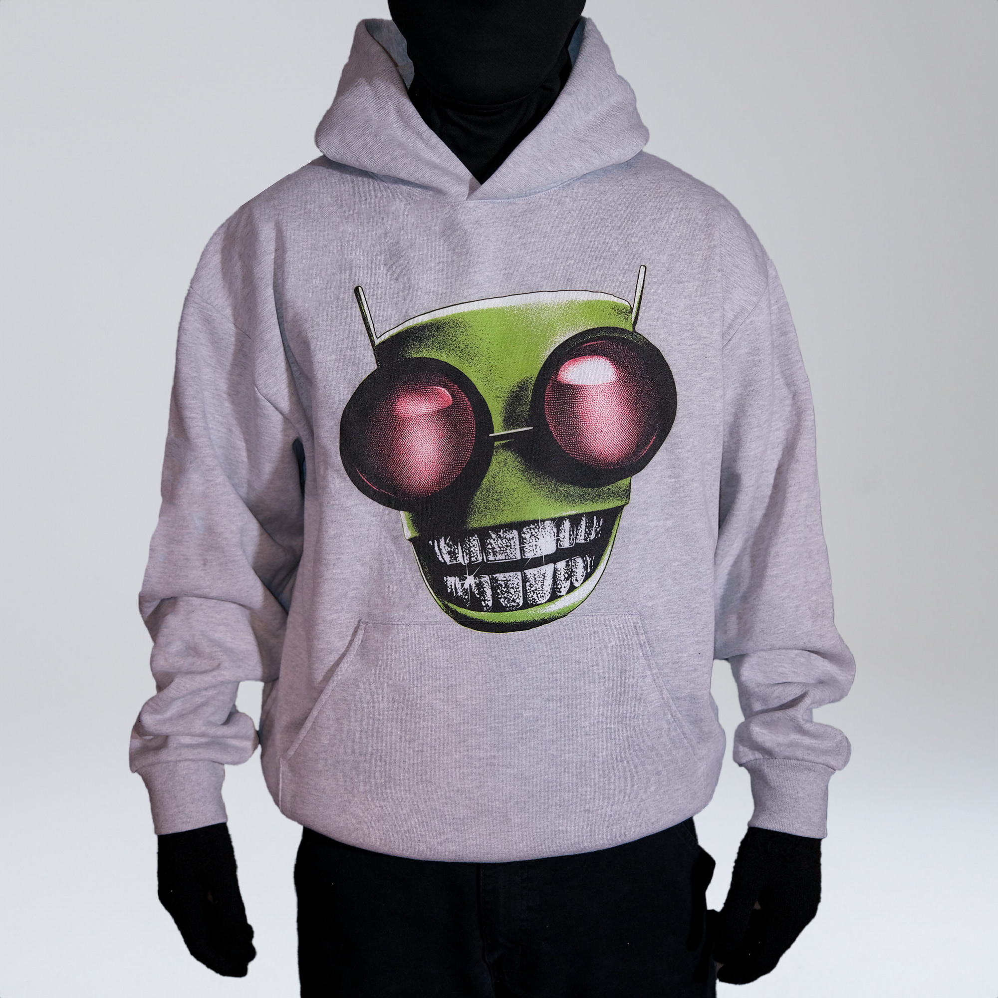 Model wearing a grey hoodie with a graphic of an green alien head featuring large red eyes and diamond grillz, standing against a neutral background. 