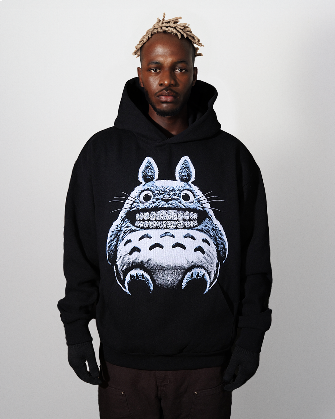 Male model wearing a black hoodie with a graphic of a light blue and tan fuzzy creature that has diamond grillz. set on a neutral background.