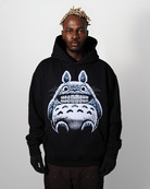 Male model wearing a black hoodie with a graphic of a light blue and tan fuzzy creature that has diamond grillz. set on a neutral background.